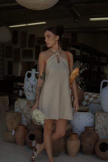 Woven dress. Adjustable halter neckline with tie closure. Back hook and eye and zipper closures. Fully lined. European summer. European summer dress. European summer aesthetic. Tuscany outfit inso. Tuscany aesthetic. Summer dress. oat mini dress. beige mini dress. tan mini dress. mini halter neck dress. summer outfit. everyday dress. vacation dress. outfit to wear to tuscany. nice dinner dress. dinner dress. day dress. 